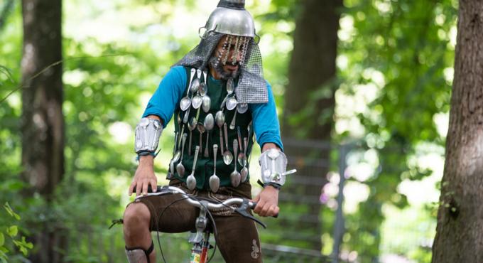 Performer in selbstgebastelter Ritterrüstung aus Löffeln und Metallhelm sitzt auf Fahrrad im Wald