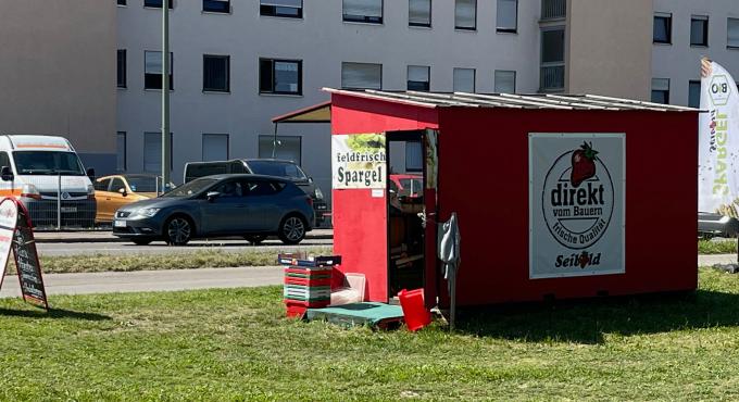 Roter Verkaufsstand von Biohof Seibold mit Schriftzug „feldfrisch Spargel" und „direkt vom Bauern" an einer Straße in Inningen.