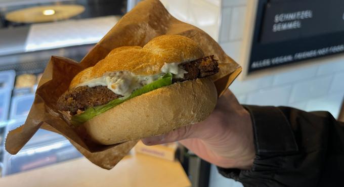 Hand hält eine vegane Schnitzelsemmel mit Salatblatt und Remoulade, im Hintergrund die Theke der Veganen Fleischerei Augsburg.
