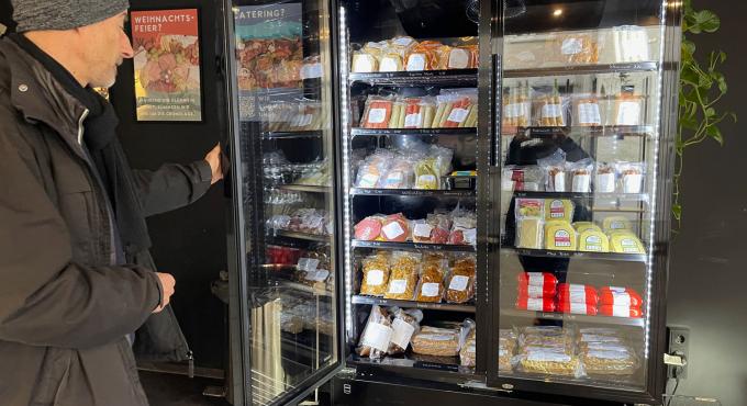 Kunde öffnet ein gefülltes Kühlregal mit vakuumverpackten veganen Wurst- und Käseprodukten in der Veganen Fleischerei Augsburg.