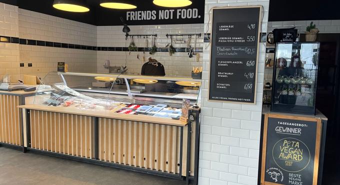 nnenansicht der Veganen Fleischerei Augsburg mit Glastheke, Kreidetafel-Speisekarte und dem Schriftzug „Friends Not Food" an der Wand.