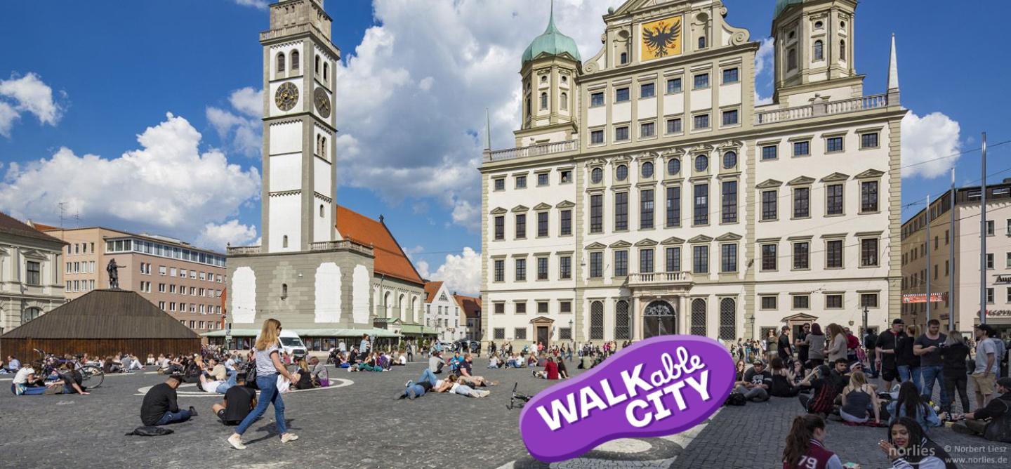 Das Bild zeigt den Rathausplatz mit Blick auf den Perlachturm und das Rathaus im Sonnenschein. Eine Frau läuft durch die Menschen, sie am Boden sitzen und die Atmosphäre genießen. Bild Norbert Liesz