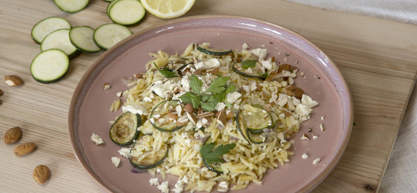 Das Bild zeigt einen Teller mit Zuchini-Zitronen-Orzo mit gerösteten Zucchinischeiben, Madelnsplittern und Flöckchen von Schafskäse bestreut. Der rosafarbene Teller steht auf einem Holzbrett auf dem links neben dem Teller Madeln, Zucchini-Scheiben und eine Zucchini sowie eine halbe Zitrone zu sehen sind. Bild Oekokiste e.V.